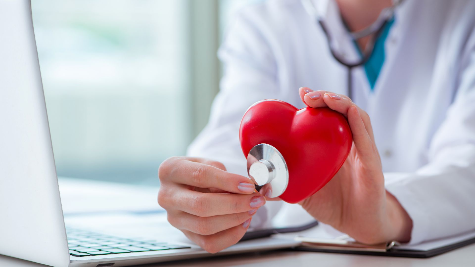 Doctor holding a red heart model and a white object near a laptop.
